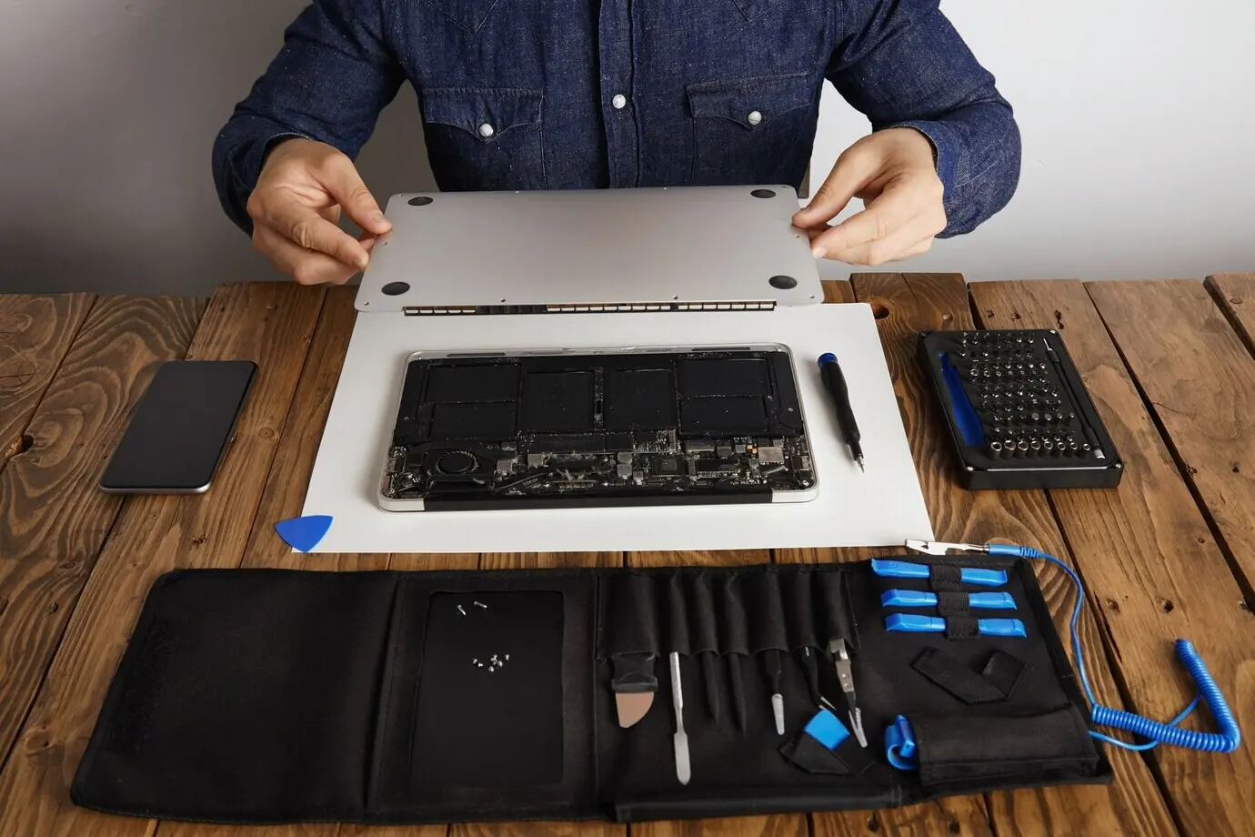 Ein Servicetechniker öffnet die rückseitige Topcase-Abdeckung eines Laptop-Computers, bevor er ihn mit seinen professionellen Werkzeugen aus der in der Nähe auf einem Holztisch befindlichen Werkzeugkiste repariert, reinigt und instand setzt; Vorderansicht.